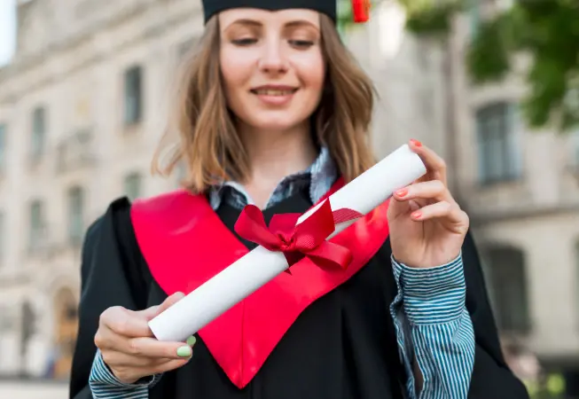 Best Online Diploma Programs in Qatar-A female graduate wearing a black academic gown and mortarboard holds a rolled diploma tied with a bright red ribbon. She stands outdoors near a university building, creating a celebratory scene that represents graduation, academic achievement, and completing a diploma program.