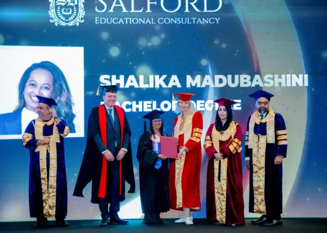 Online Bachelor’s Degrees in Qatar-A group of graduates and academic officials pose on stage during a formal master’s degree ceremony, wearing colorful graduation gowns and caps. One graduate proudly holds a certificate, with a large LED screen displaying the event title in the background.