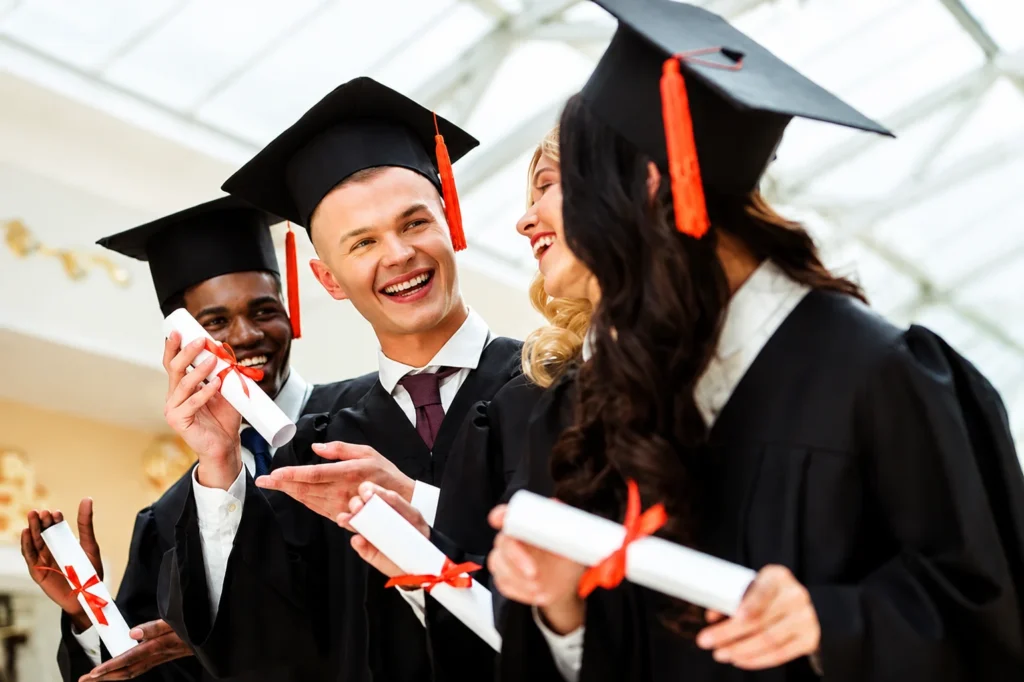 UK Diploma Levels in Qatar -A group of diverse university graduates are celebrating together in a bright indoor setting. They are wearing black graduation gowns and mortarboards with red tassels, while proudly holding rolled diplomas tied with red ribbons. The students are smiling and interacting naturally, creating a joyful and successful graduation atmosphere.